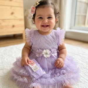 Smiling baby girl wearing a purple tulle party dress with floral details sitting on a soft rug indoors